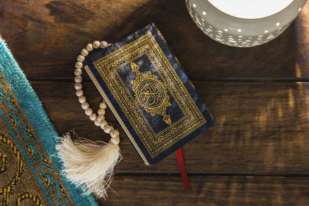 Al-Qur’an berwarna biru dengan hiasan emas di atas meja kayu, disertai tasbih dan cahaya lampu.
